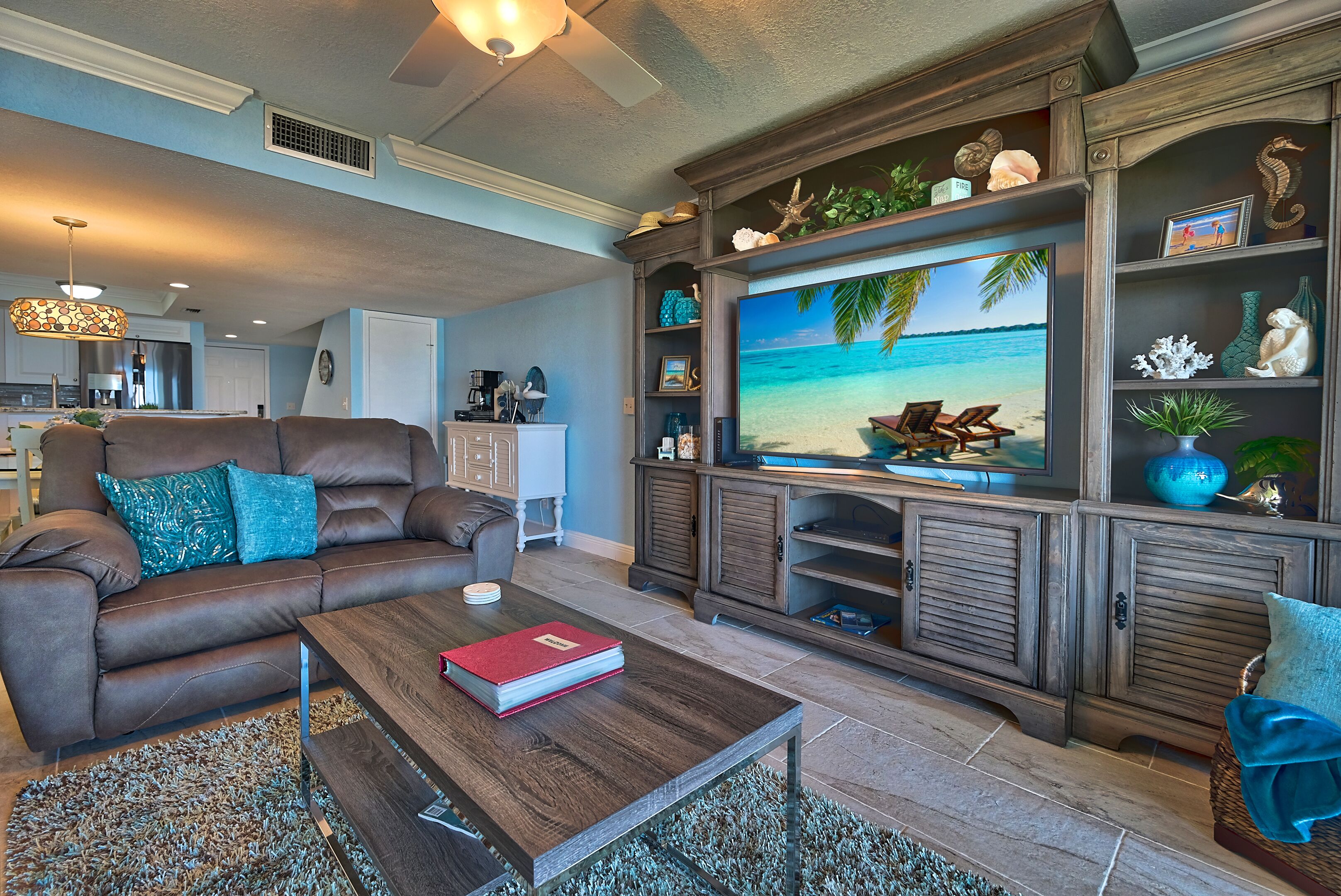 Un soggiorno con un divano in pelle marrone, un tavolino da caffè con un libro, una televisione che mostra una scena di spiaggia e un mobiletto in legno con oggetti decorativi.
