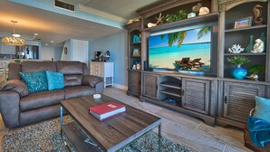 Una sala de estar con un sofá de cuero marrón, una mesa de centro con un libro, un televisor mostrando una escena de playa y un mueble de madera con objetos decorativos.