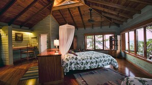 A bedroom with a wooden ceiling, a bed with a floral bedspread, a nightstand, a desk with a chair, and a window with a view of the outdoors. - Mariposa Del Mar: Spectacular Ocean front Villa with private pool. (Roatan)