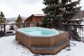 Un bain à remous en bois entouré de neige, avec une rambarde en métal et une maison en arrière-plan.