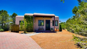 A single-story house with a covered porch, a front door, and a small garden area with plants and a bench. - Peaceful Hilltop Casita w/ Stunning Views, Close to the Historic Plaza (Santa Fe)