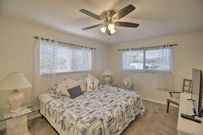 A bedroom with a bed, two bedside tables, a ceiling fan, a chair, a desk with a computer, and two windows with blinds.