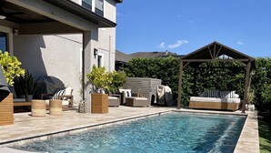 Une piscine dotée de chaises longues et d’un espace salon couvert, le tout entouré d’une maison et de verdure.