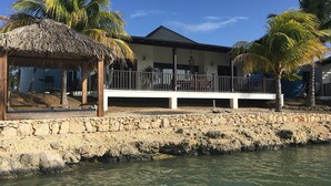 Una propiedad frente a la playa con una estructura de techo de paja, un balcón blanco y un muro de piedra.