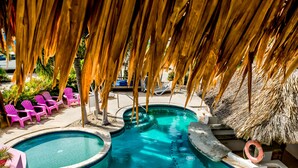 Un área de piscina con dos piscinas separadas, sillones de descanso y una estructura de techo de paja.