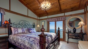 A bedroom with a large bed, wooden ceiling beams, a chandelier, a dresser, and a view of the outdoors. - Oceanfront Vacation Home - Spectacular Views; Hot Tub; Wi-Fi. (Elk)