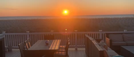 Une terrasse en bois, dotée de mobilier d’extérieur, d’une rambarde et offrant une vue sur le coucher de soleil sur l’océan.
