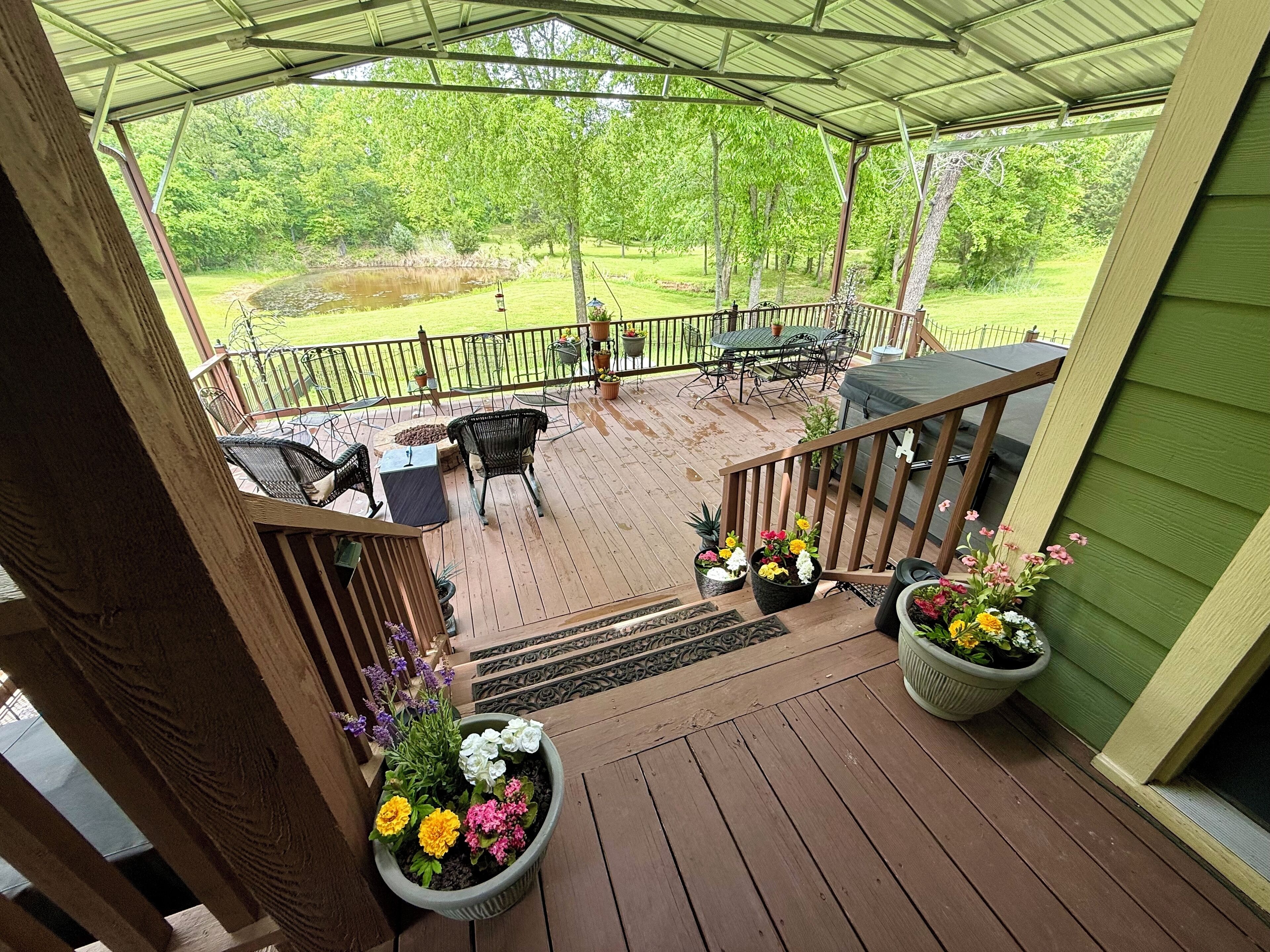 Eine überdachte Terrasse im Freien mit Holzboden, umgeben von einem Metallgeländer. Mehrere Blumentöpfe und ein Tisch mit Stühlen sind vorhanden.