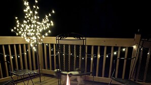 Een vuurplaats met een stenen rand op een houten terras, omringd door stoelen en lichtslingers.