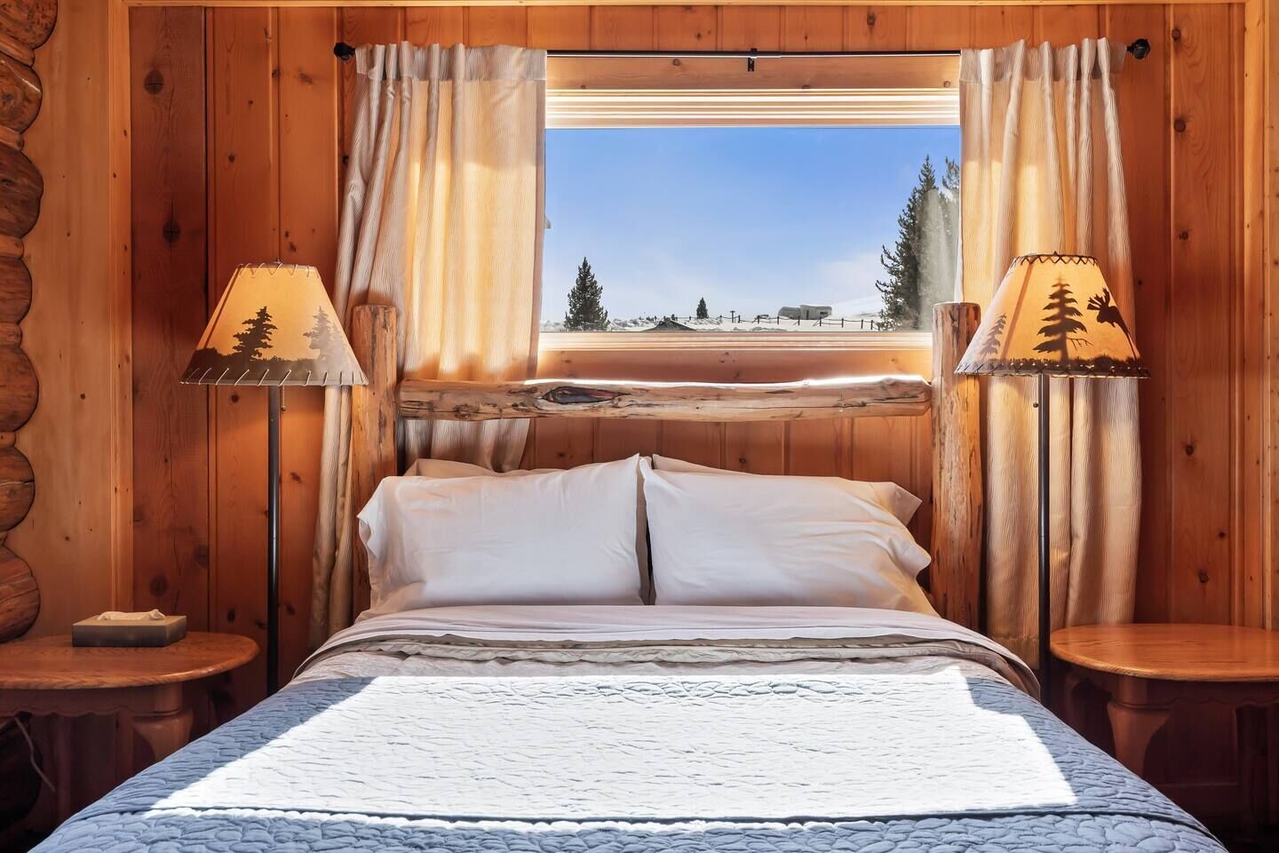 A wooden cabin bedroom with a bed, two bedside lamps, and a window with a scenic view.