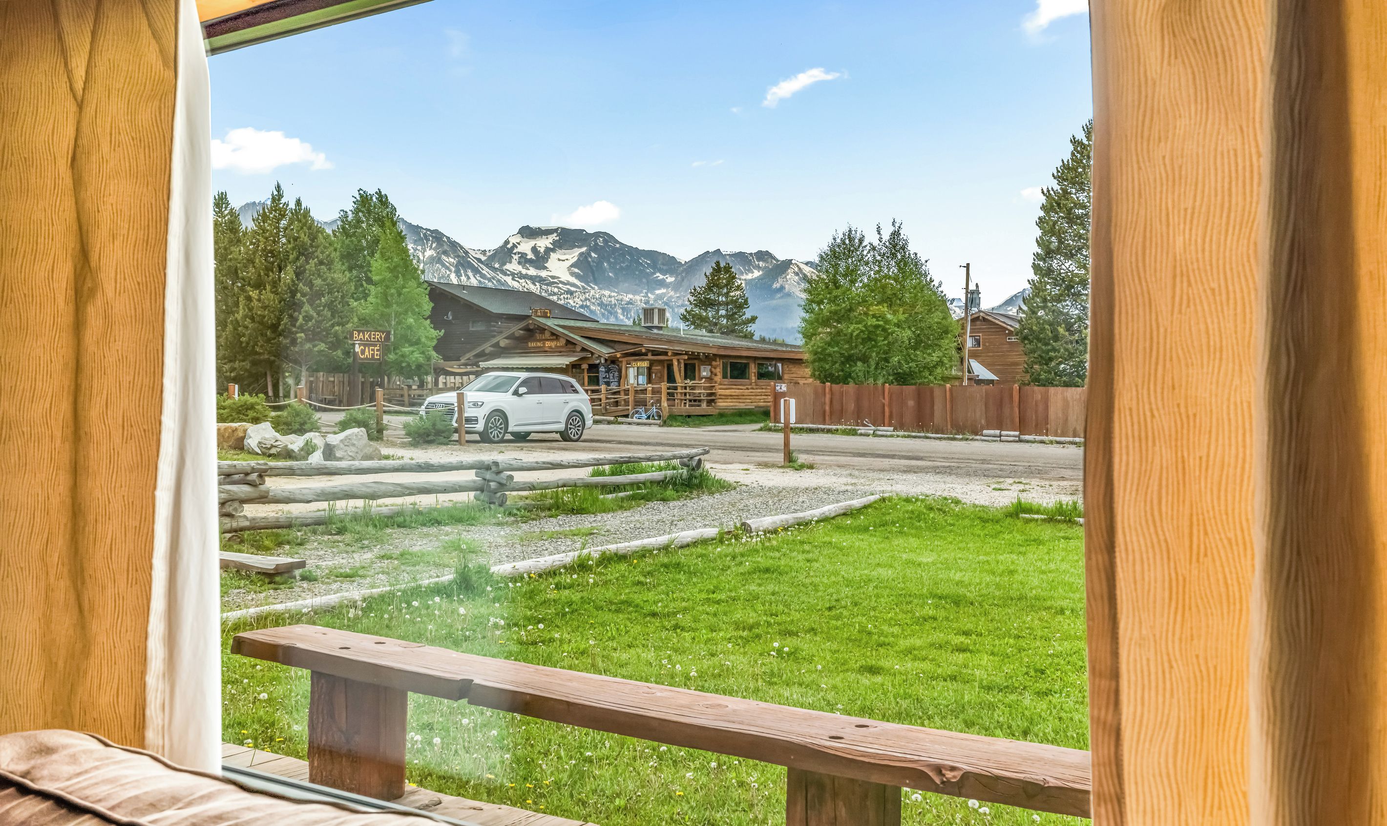 A view from a window overlooking a wooden building with a sign for a bakery cafe, a white car parked in front, and a mountain backdrop.