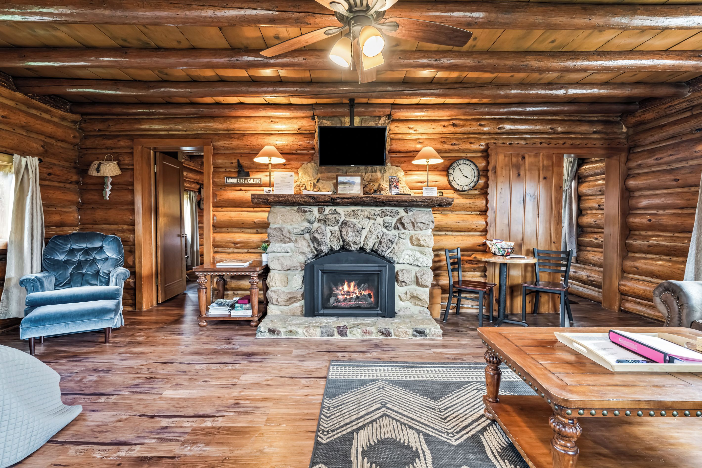 L’intérieur d’une cabane en rondins, avec une cheminée en pierre, des meubles en bois et un ventilateur de plafond.