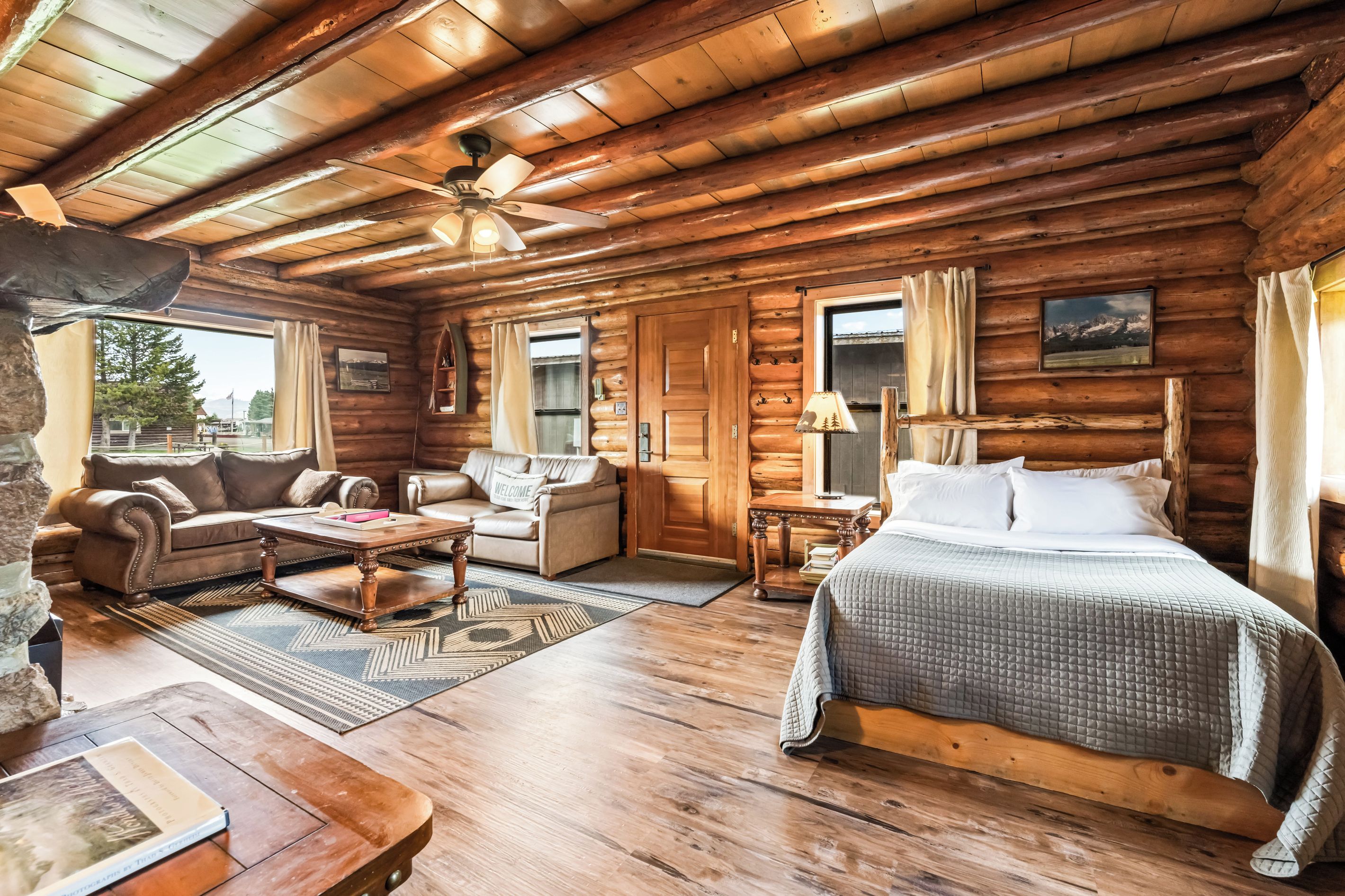 Interior de uma cabana de tronco com uma sala de estar, sofá, mesa de centro, cama e janela com cortinas.