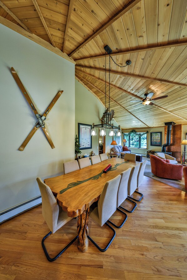 A spacious dining area with a long wooden table and cushioned chairs, a wooden ceiling with exposed beams, and a hanging light fixture.