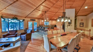 Un vaste espace dédié à la salle à manger, avec une grande table et des chaises en bois, une cuisine à l’arrière-plan et une vue sur la forêt environnante grâce à de grandes fenêtres.
