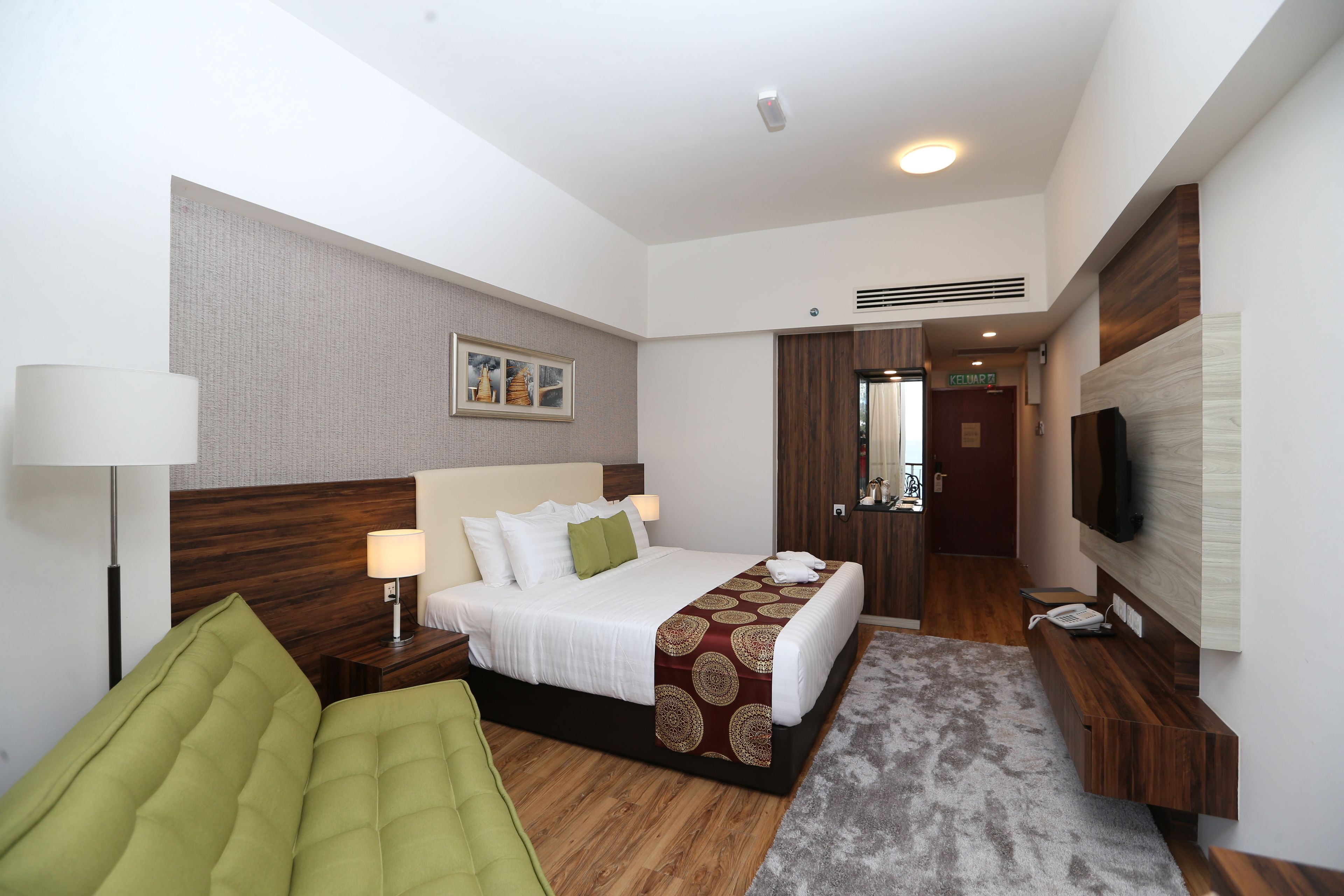 A modern hotel room with a large bed, a green armchair, a desk with a telephone, and a flat-screen TV mounted on the wall.