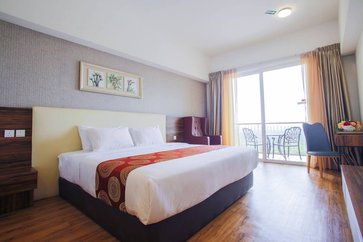 A hotel room with a large bed, a wooden nightstand, a desk with a chair, and a balcony with a table and chairs.