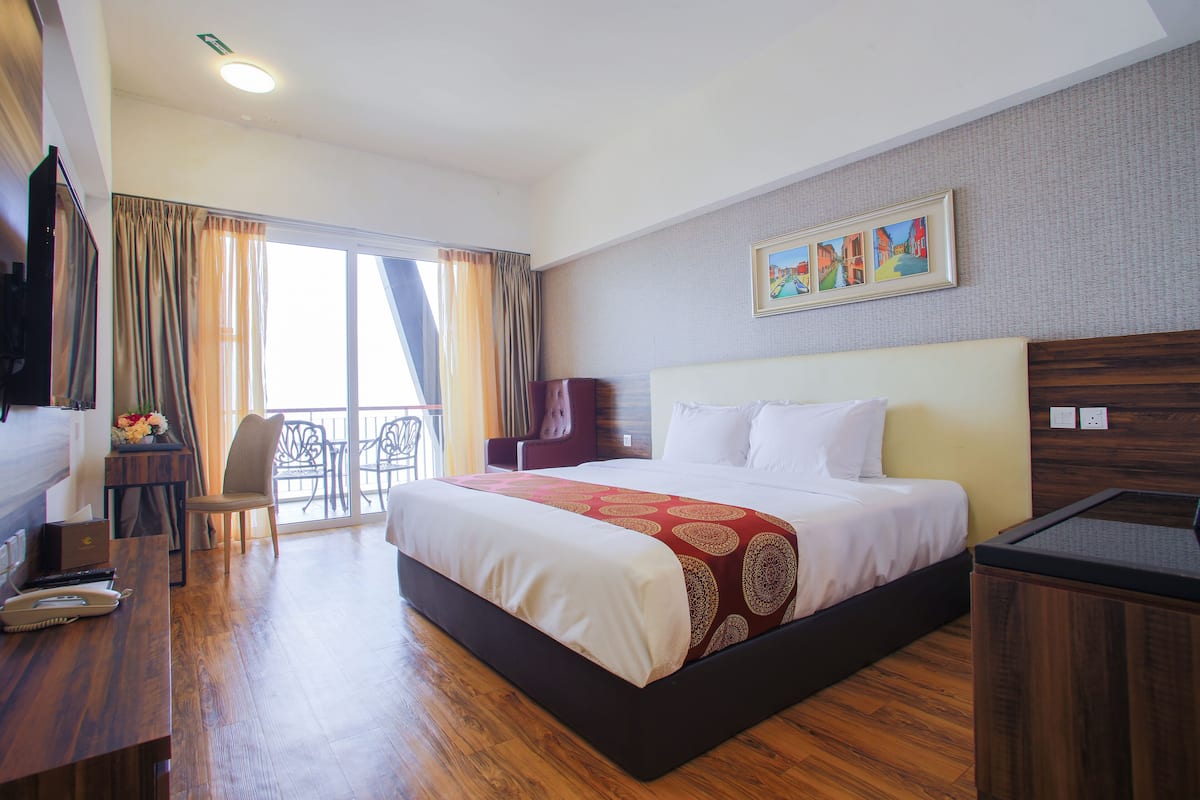 A hotel room with a large bed, a desk with a laptop, a chair, and a view of the outdoors through a large window.