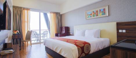 A hotel room with a large bed, a desk with a laptop, a chair, and a view of the outdoors through a large window.
