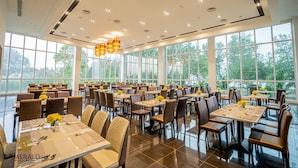 A spacious dining area with multiple tables and chairs, large windows offering a view of trees, and modern pendant lighting. - Amerald Resort Hotel Desaru (Bandar Penawar)