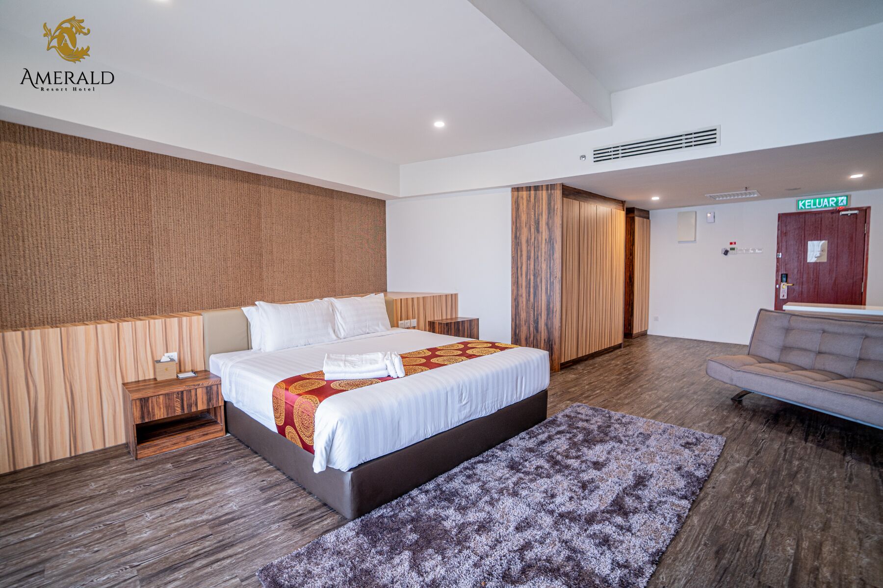 A hotel room with a large bed, wooden headboard, a grey sofa, and a patterned rug.
