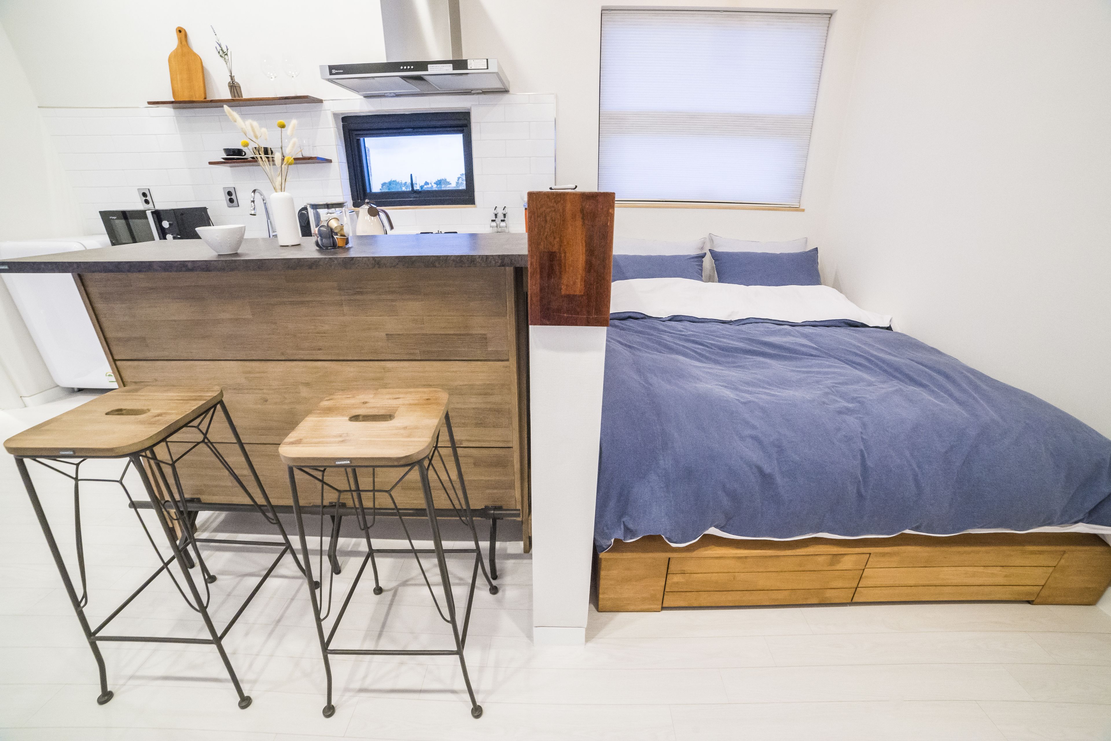 Un dormitorio acogedor con cama, cabecera de madera y pequeña zona de kitchenette con taburete y ventana.