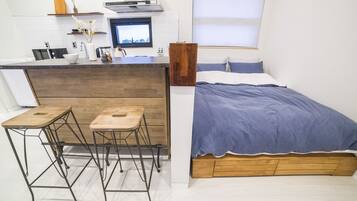 Un dormitorio acogedor con cama, cabecera de madera y pequeña zona de kitchenette con taburete y ventana.