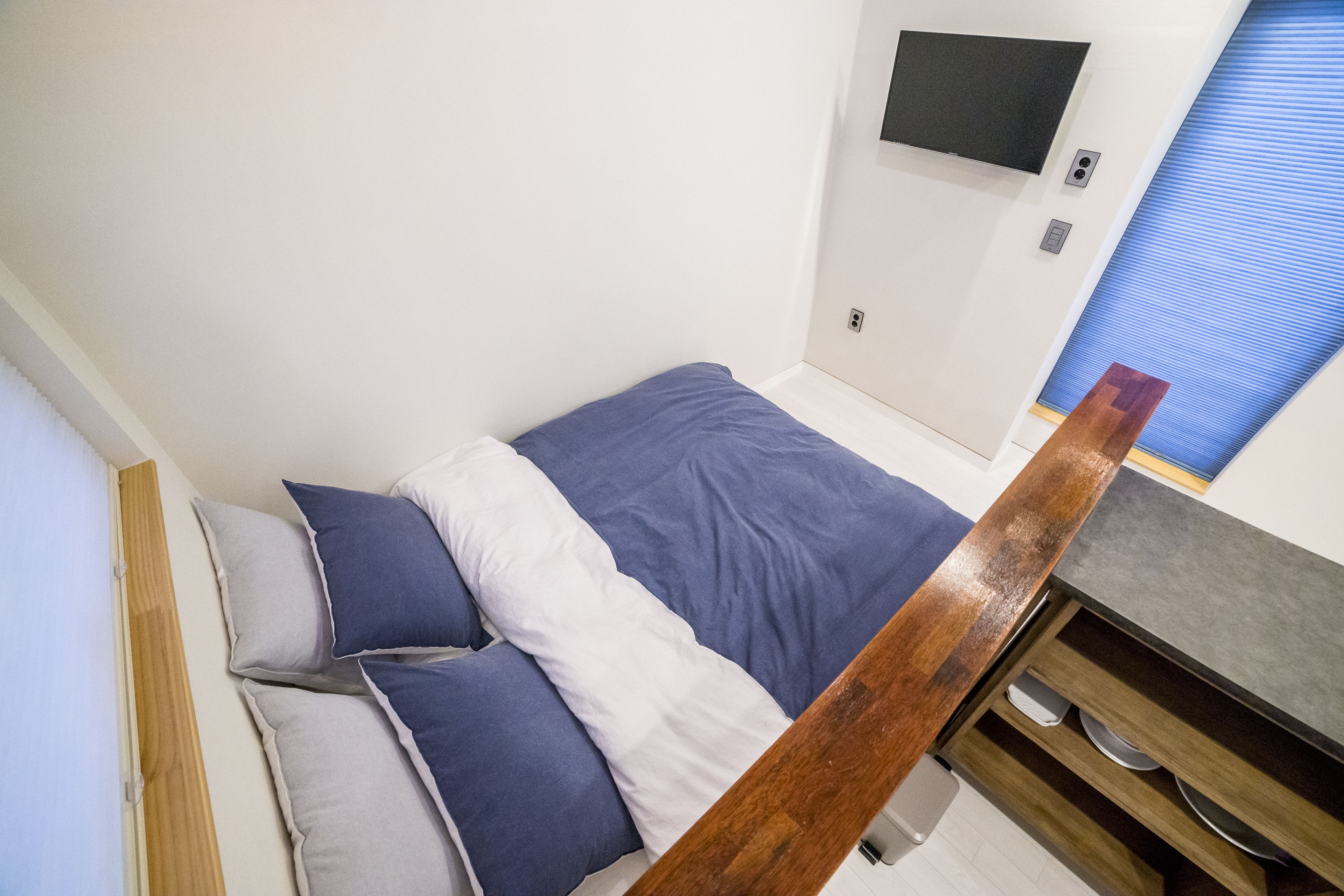 Ein Schlafzimmer mit Bett, Kissen, einem Holzkopfstück, einem an der Wand montierten Fernseher und einem Fenster mit Jalousien.