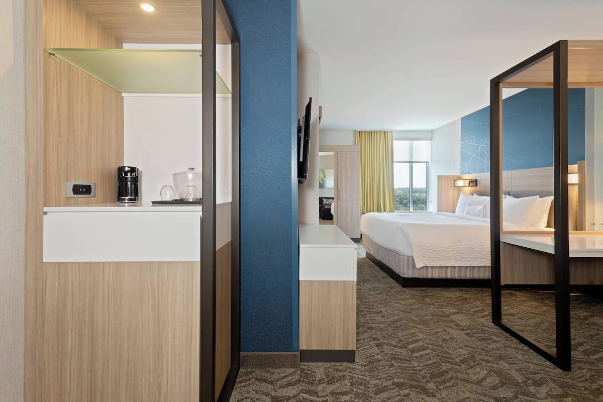 A hotel room with a bed, bedside table, and a mirror. There is a window with curtains and a view of the outside.