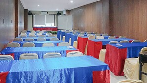 Une salle de conférence avec des rangées de tables recouvertes de nappes bleues et rouges, des chaises beiges et un grand écran à l’avant.
