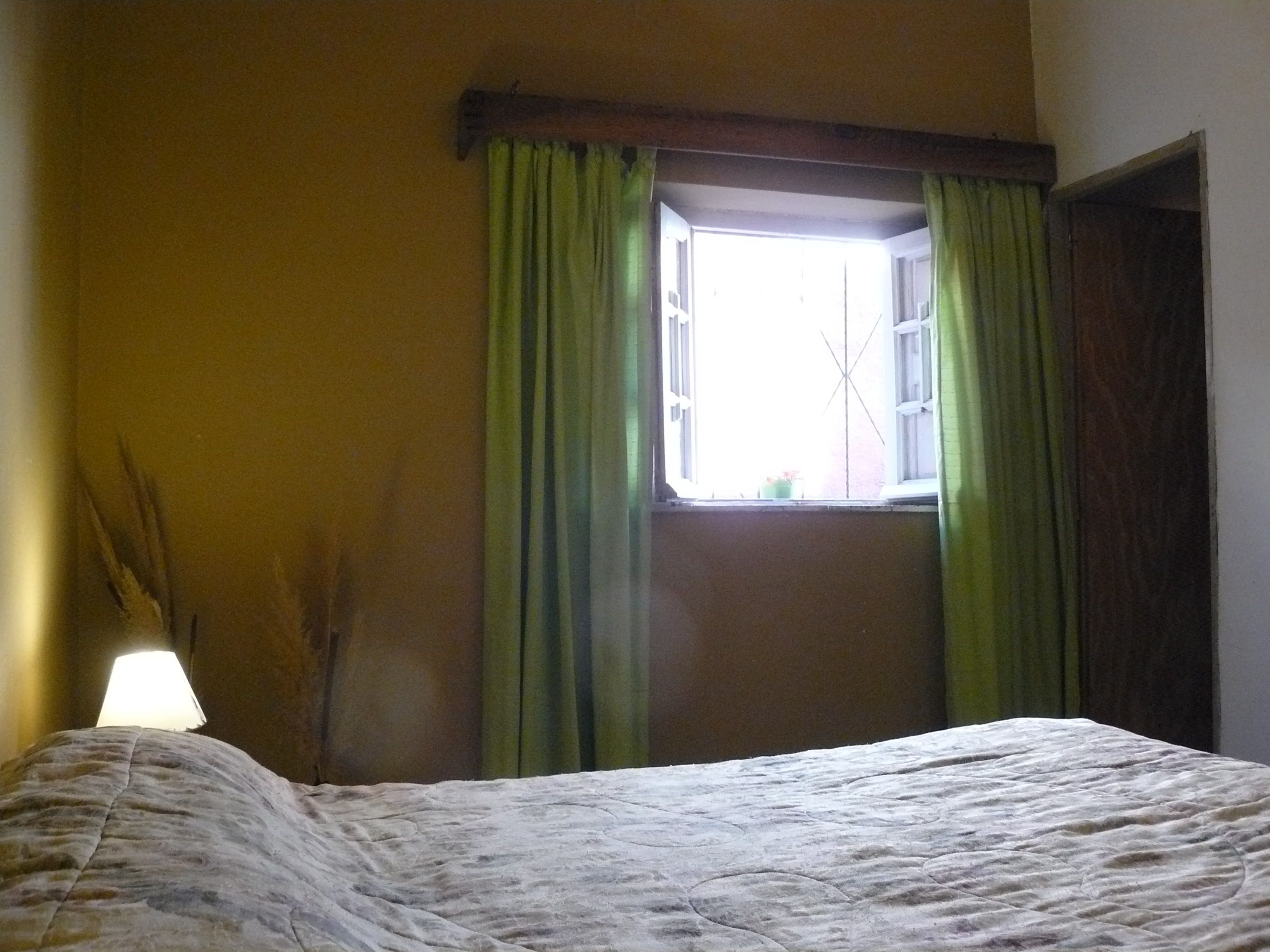 Una habitación con una cama, un ventanal con cortinas verdes y una lámpara sobre una mesita de noche.