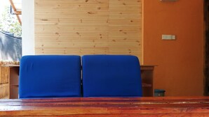 A room with a wooden ceiling, two blue chairs, a wooden table, and an orange wall with a light switch. - Foret De Vill - Hostel (Ella)