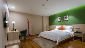 A modern hotel room with a wooden floor, a bed with white and orange pillows, a desk with a lamp, a green chair, and a small table with a plant. - Sucha Hotel Xian Bell Tower Huimin St (Xi'an)
