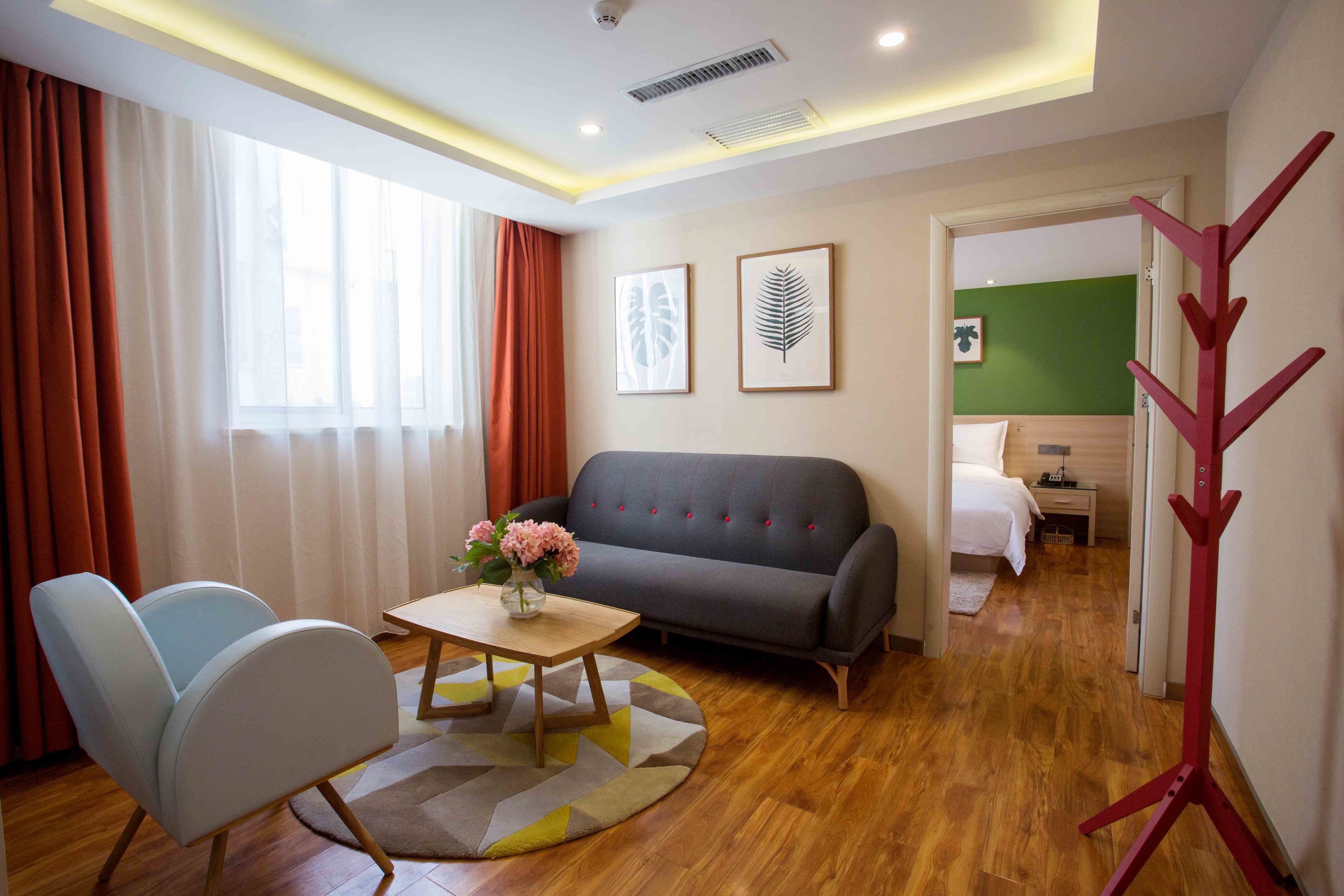 Habitació d'hotel moderna amb un sofà gris, una taula de centre de fusta amb un ram de flors, un butacó blau i un llit amb roba de llit blanca.