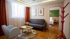 A modern hotel room with a grey sofa, a wooden coffee table with a vase of flowers, a blue armchair, and a bed with white bedding. - Sucha Hotel Xian Bell Tower Huimin St (Xi'an)
