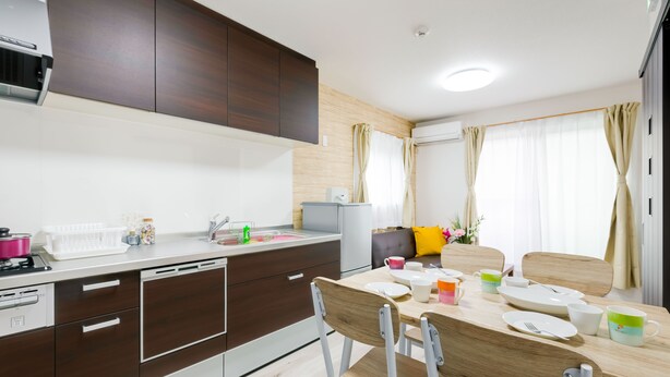 A modern kitchen with dark wooden cabinets, a white countertop, and a dining area with a wooden table and chairs. - Mikazuki House Teradacho (Osaka)
