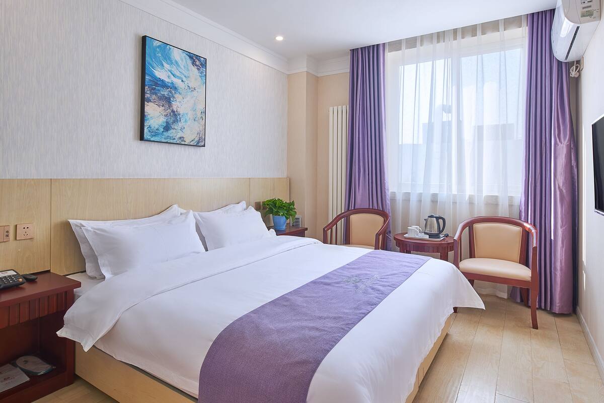 A hotel room with a large bed, a bedside table with a phone, a small table with a kettle, two chairs, and a TV mounted on the wall.