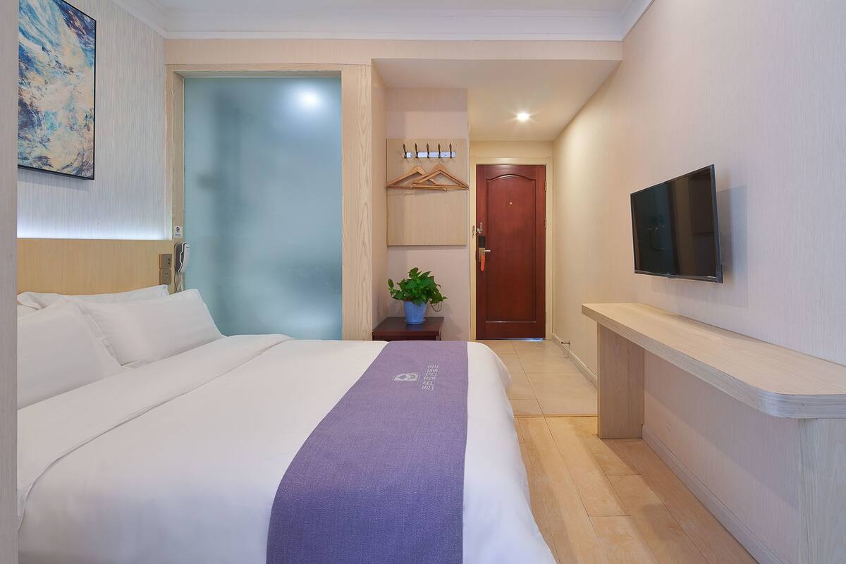 A hotel room with a bed, a TV mounted on the wall, a wooden bench, and a door leading to another room.