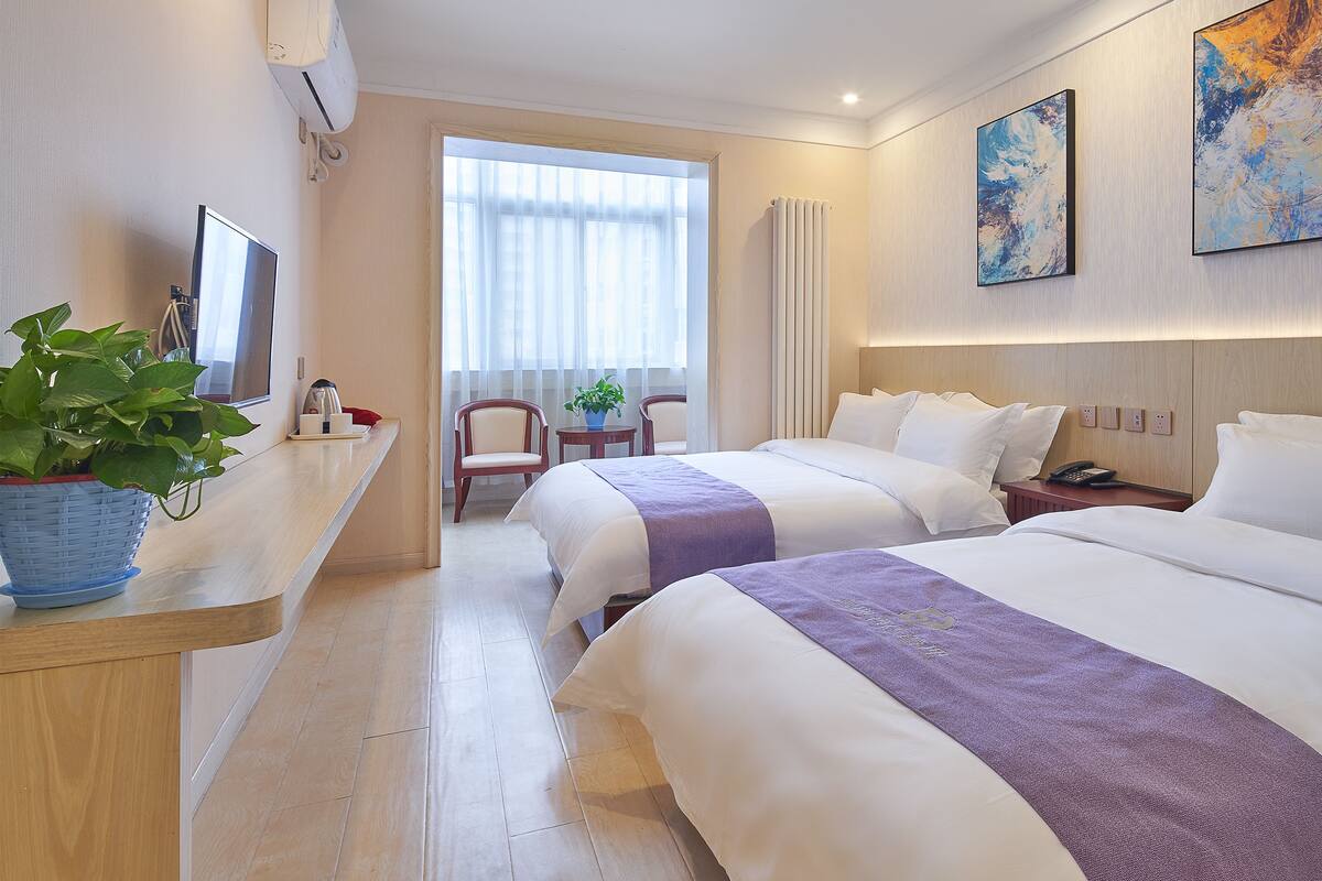 A hotel room with two beds, a desk with a plant, a chair, a window with curtains, and artwork on the wall.