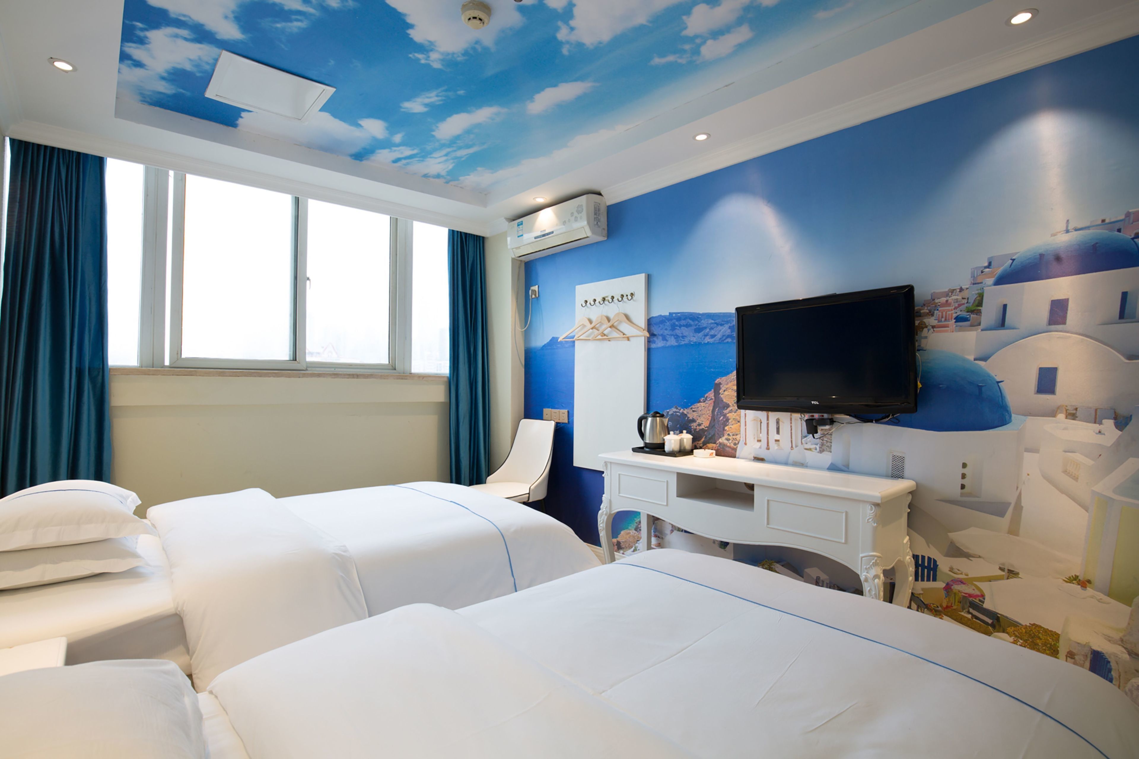 Une chambre d’hôtel avec deux lits, un bureau doté d’un téléviseur à écran plat, et une fresque représentant un ciel bleu et des bâtiments blancs.