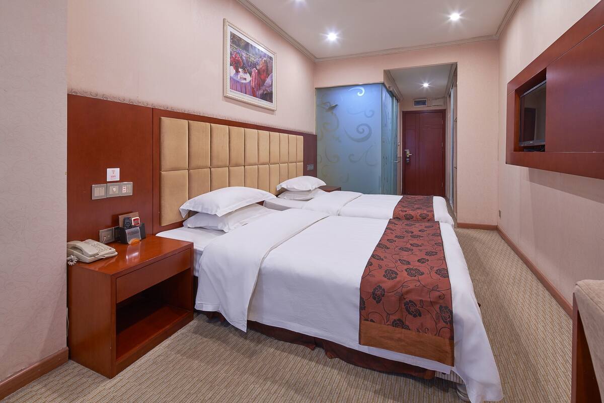 A hotel room with a large bed, a bedside table with a phone, a decorative wall hanging, and a door leading to another room.