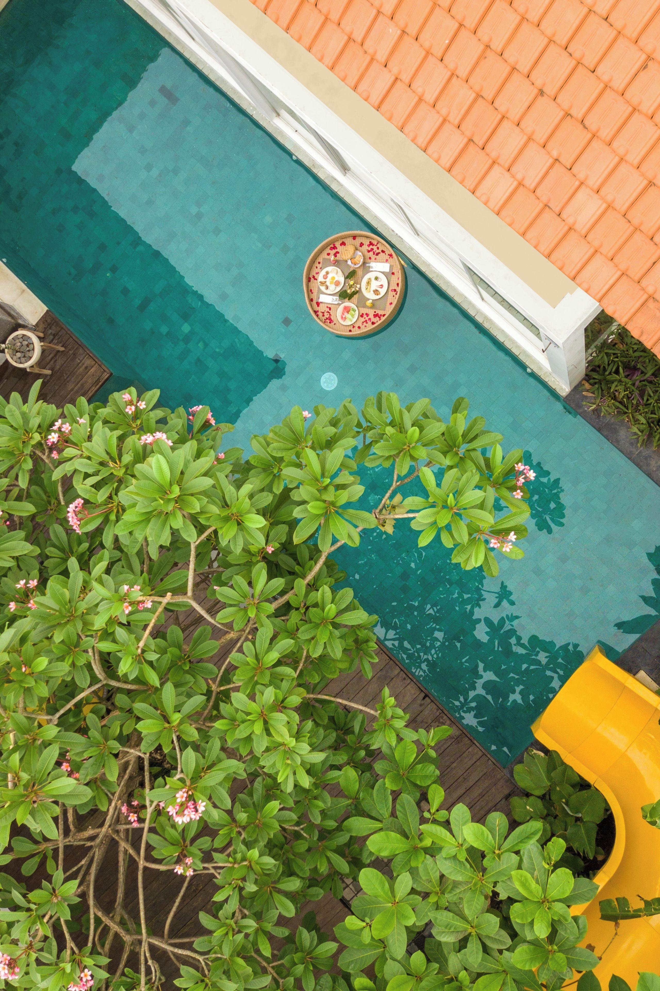 A swimming pool with a circular table in the center, surrounded by lush greenery and a yellow slide.
