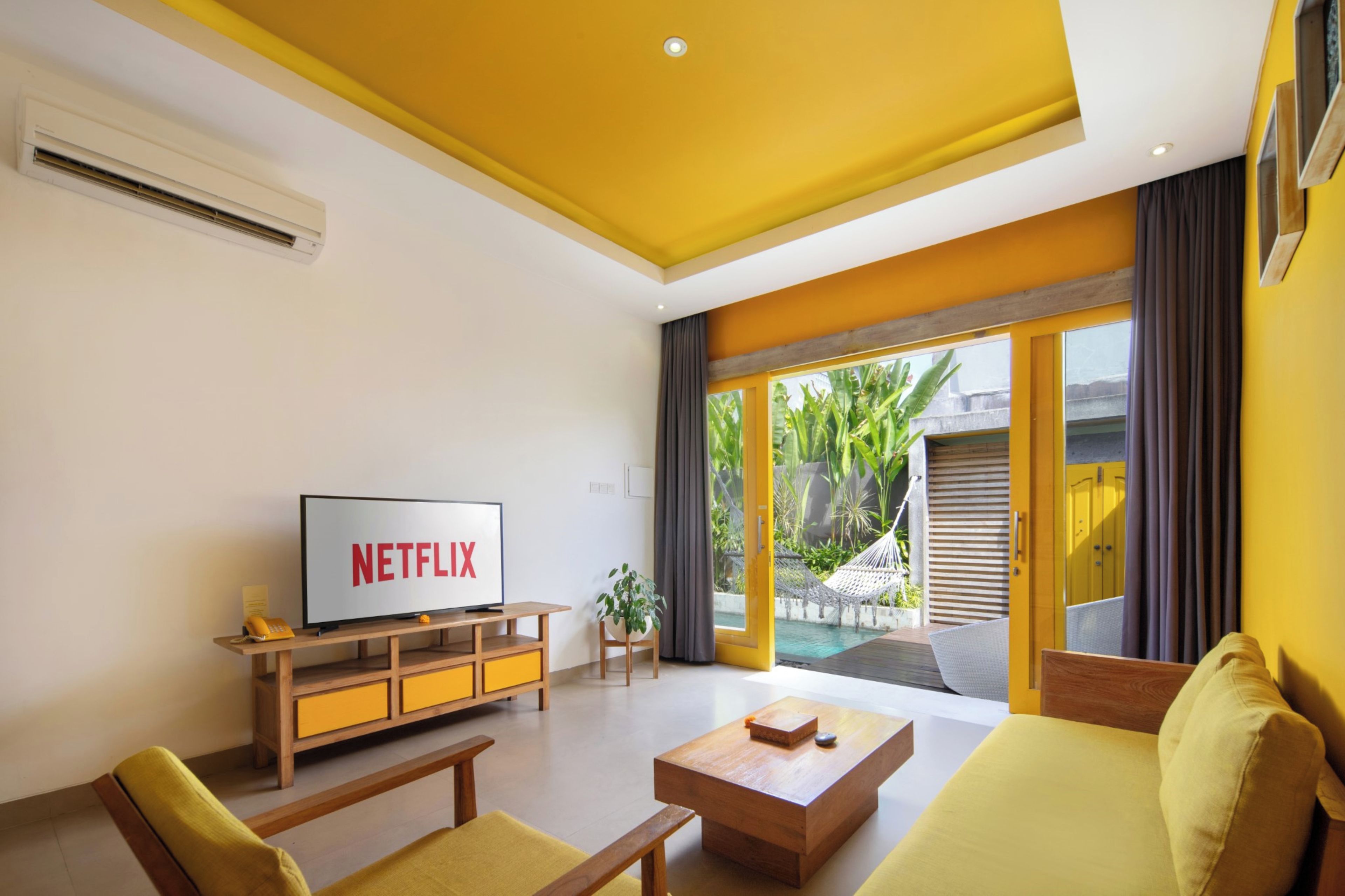Ein modernes Wohnzimmer mit einem Fernseher, der 'NETFLIX' zeigt, einem Holztisch, einer Couch und Blick auf einen Pool und Außenbereich.