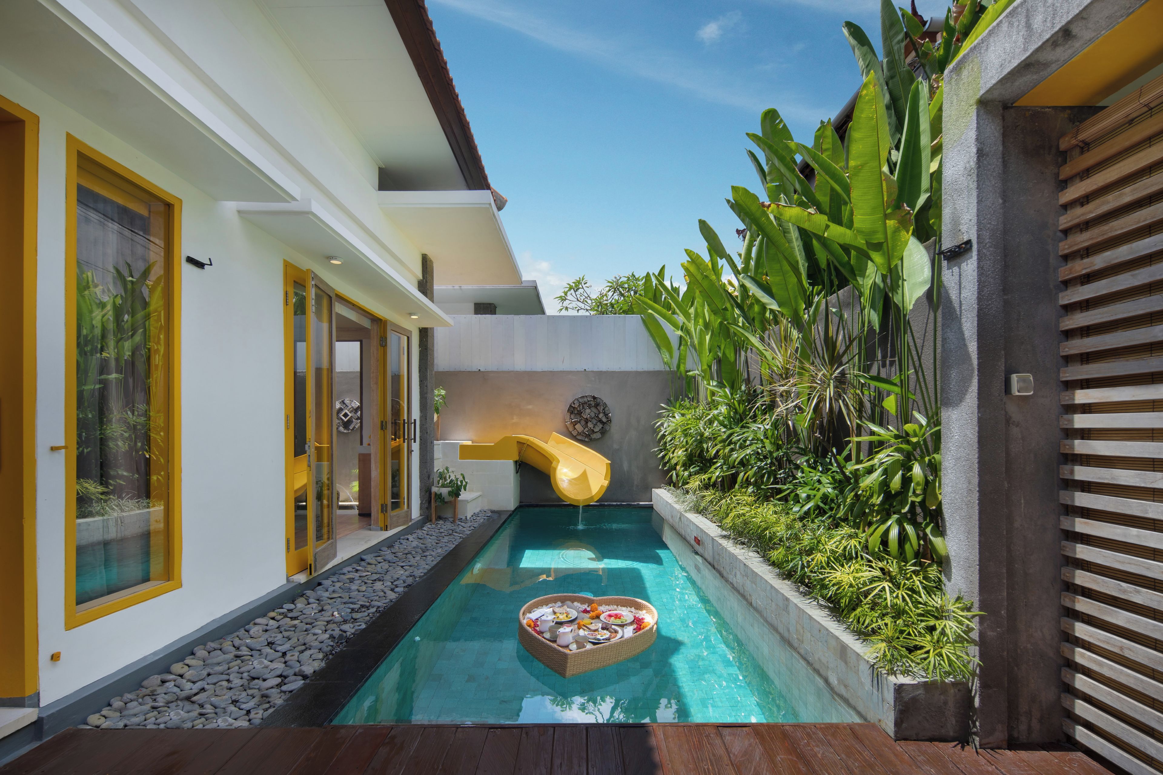 A swimming pool with a slide, surrounded by tropical plants and a wooden deck.