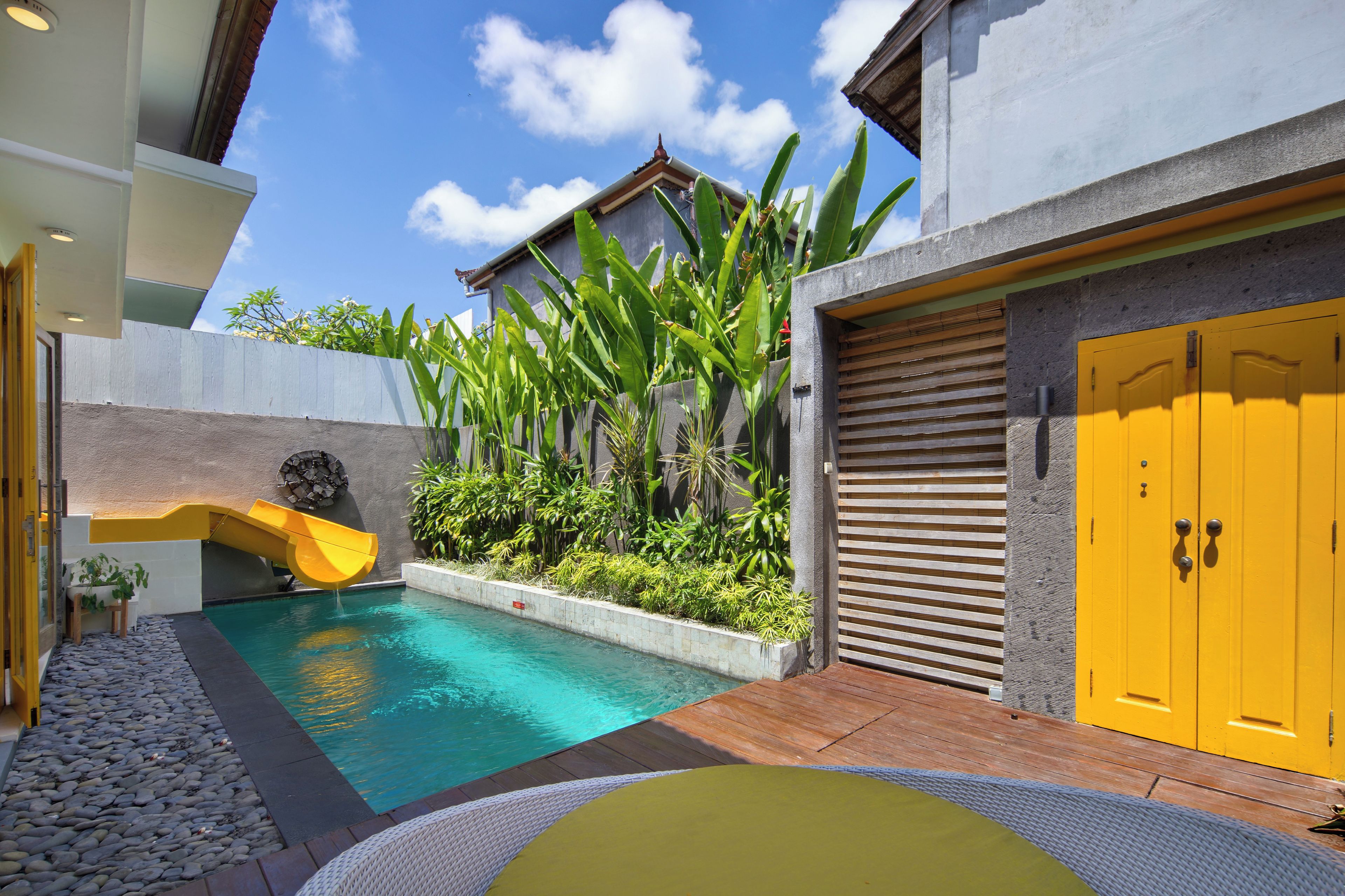 A modern outdoor pool area with a slide, surrounded by lush greenery and a yellow door.