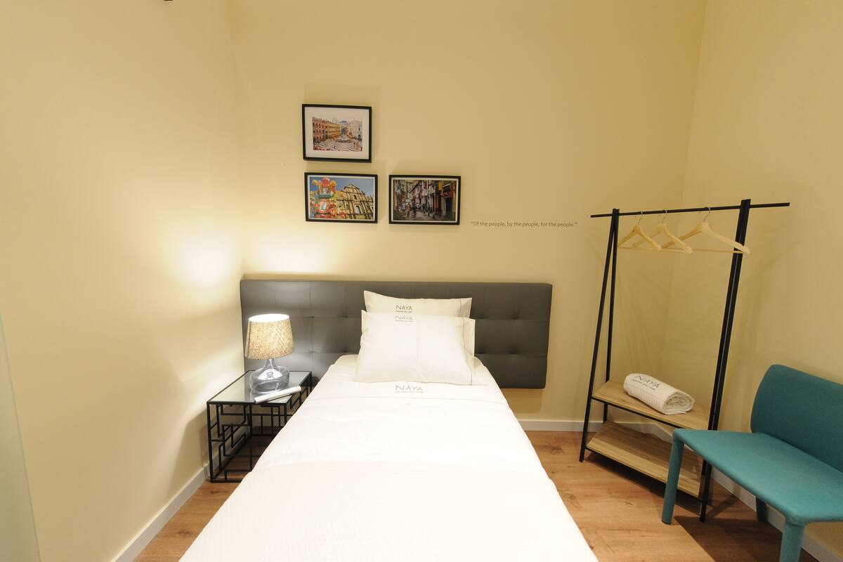 Habitació d'hotel ben ordenada amb un llit, una taula de nit amb una làmpada, una panxa i un penjador.