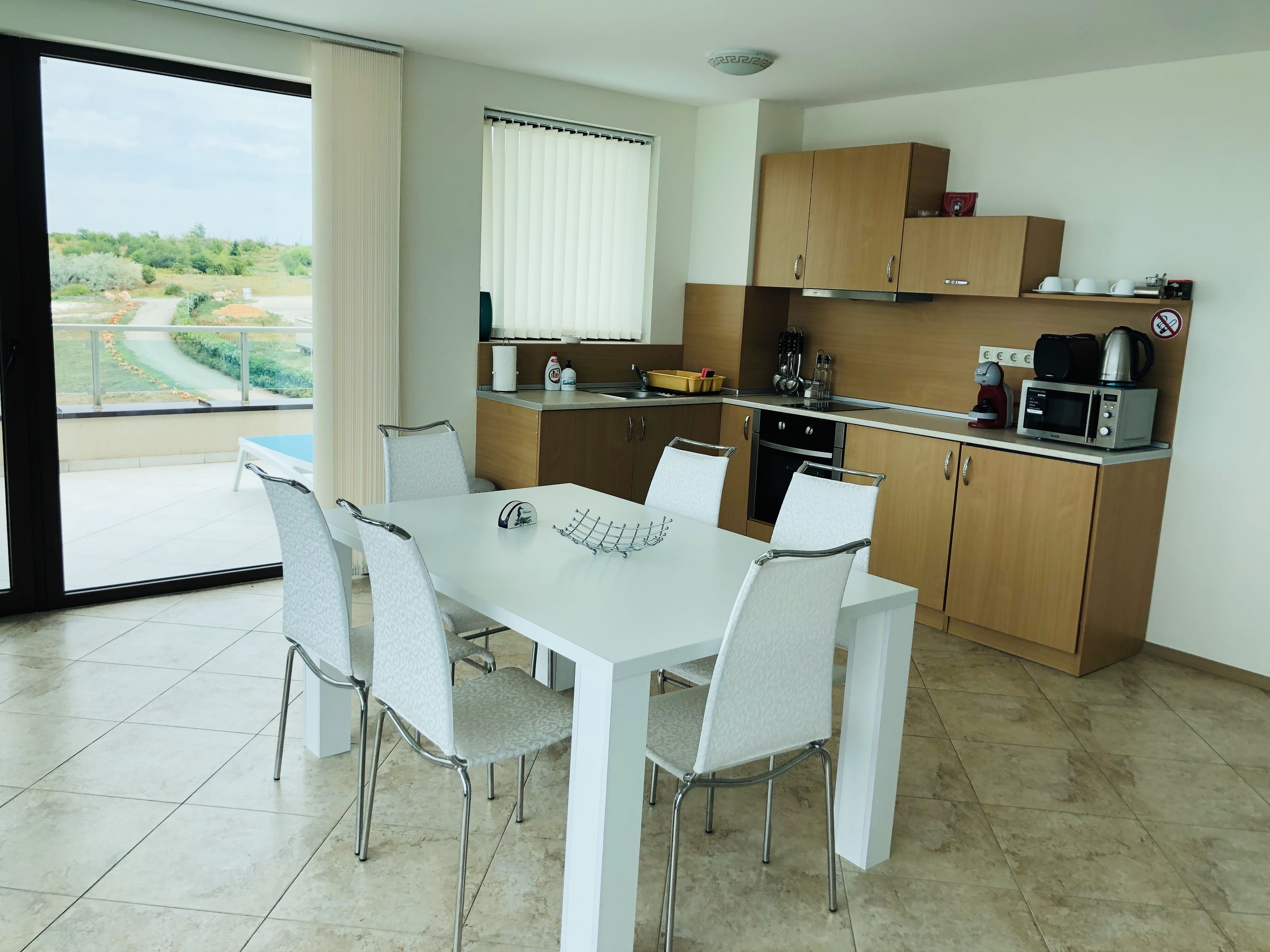 A modern kitchen with white cabinets, a dining table with four chairs, and a view of greenery outside.