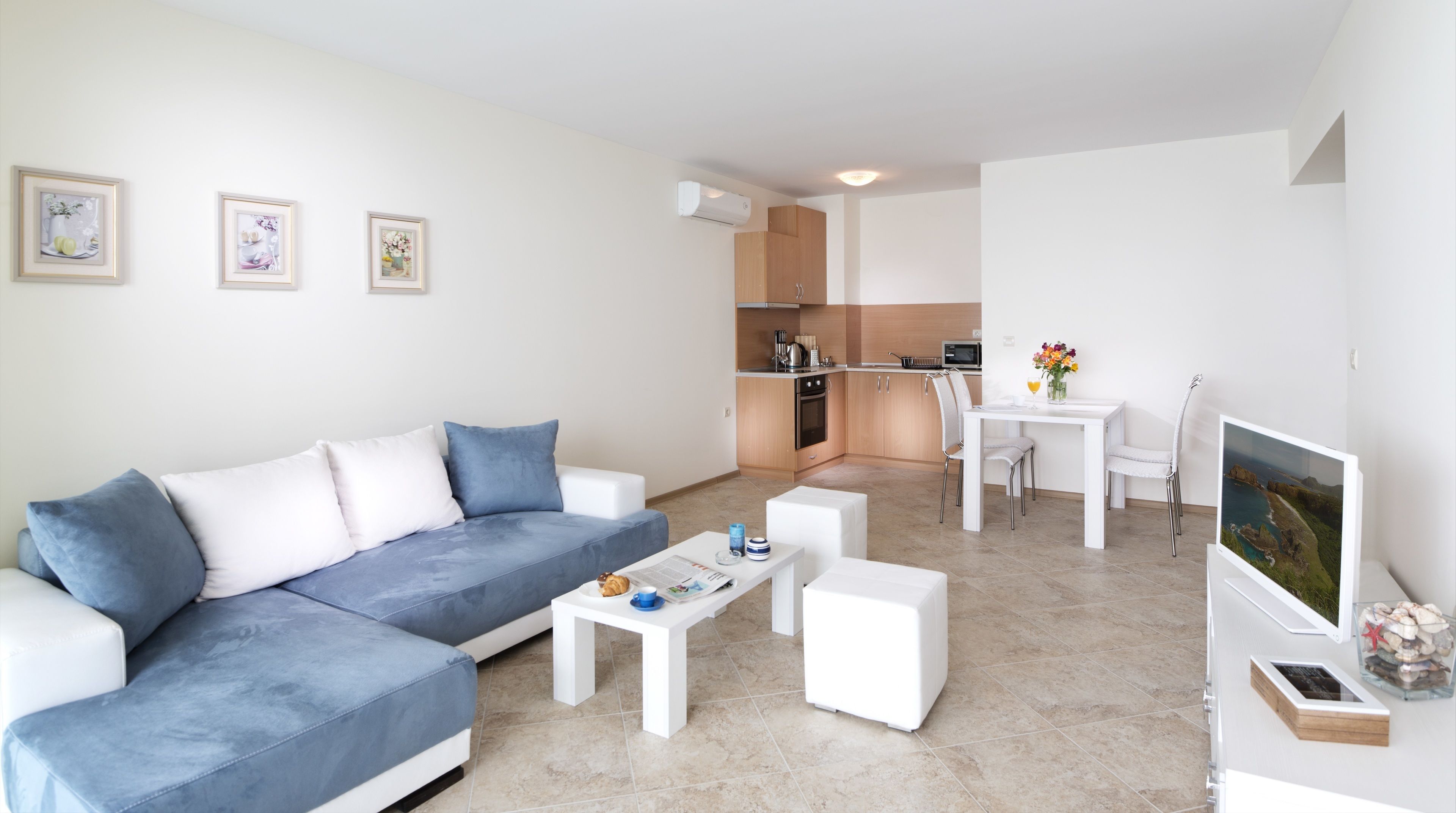 Ein modernes Wohnzimmer mit grauem Sofa, Couchtisch und Essbereich mit weißem Tisch und Stühlen. Im Hintergrund ist eine Küche mit Holzschränken und einer Mikrowelle zu sehen.