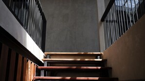 Uma escadaria com degraus de madeira e corrimãos de metal, subindo em um edifício com parede lisa e uma janela ao fundo.