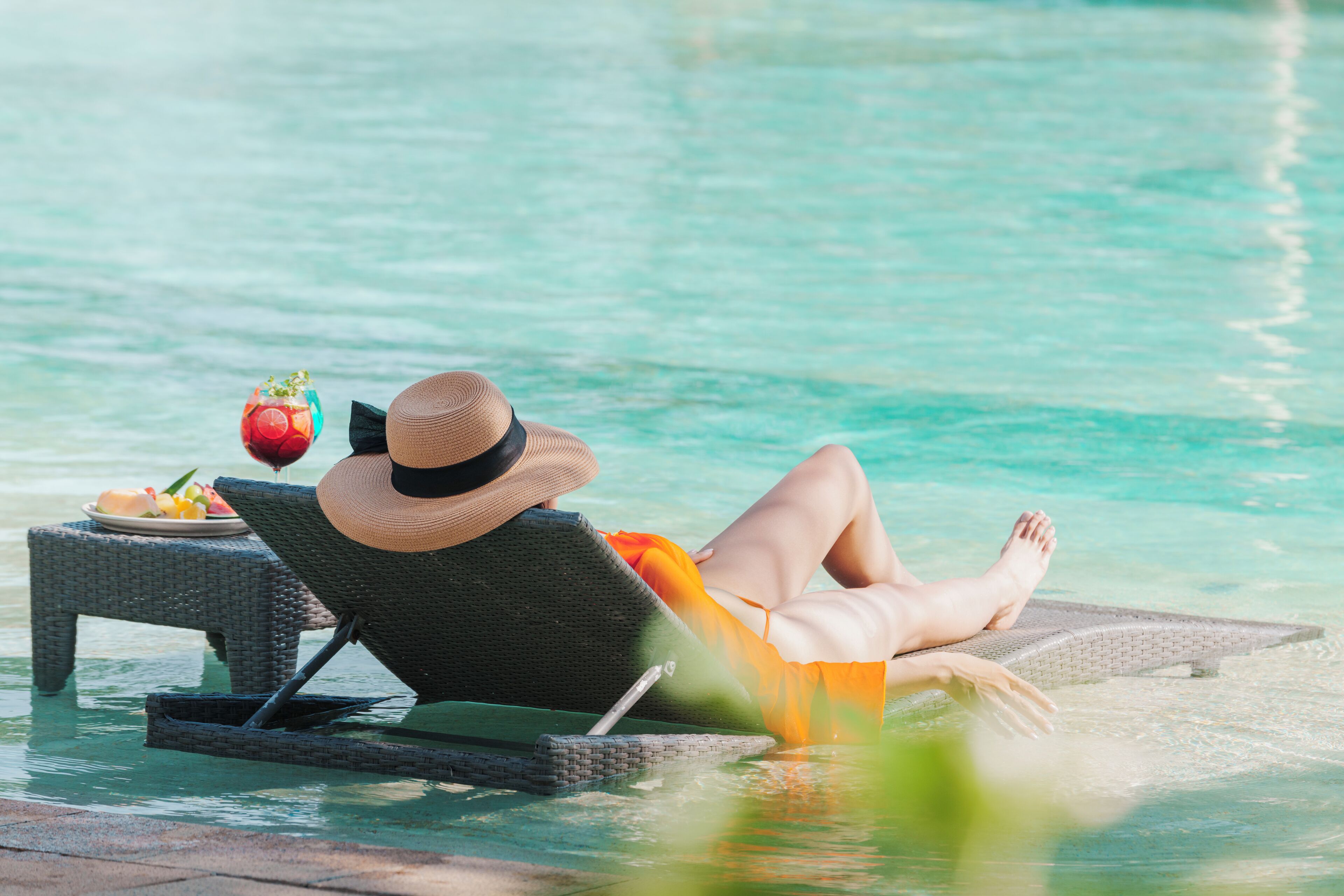 A person reclining on a lounge chair by a pool with a plate of fruit and a drink.